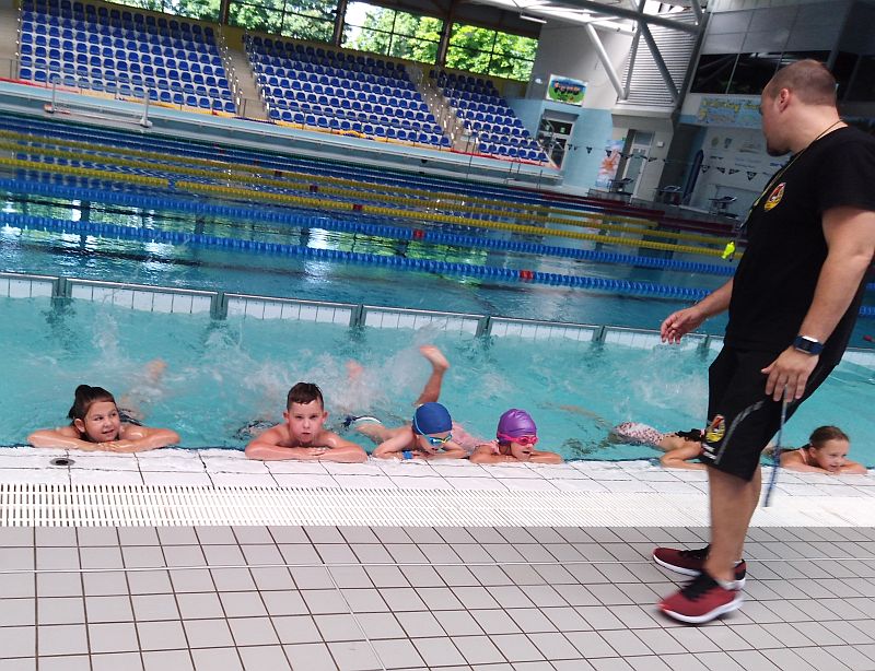 Grupa dzieci ucząca się pływać pod nadzorem instruktora stojącego na brzegu basenu